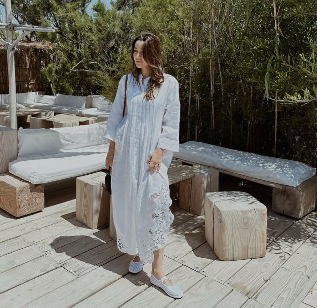 Chic woman dressed in Bearice .b in a flowing white embroidered linen dress, styled for a relaxed summer day in a serene, nature-inspired outdoor lounge setting.