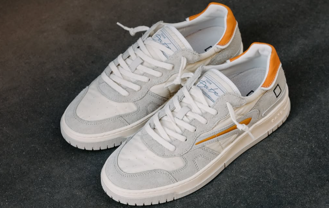Stylish pair of premium D.A.T.E. sneakers featuring a white and light gray suede upper with orange heel accents and signature branding on the tongue.