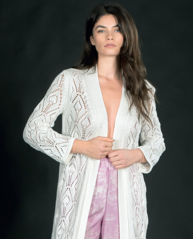 Confident woman wearing a white open-knit cardigan from Ella Lago di Como with intricate geometric patterns over soft lilac satin trousers.